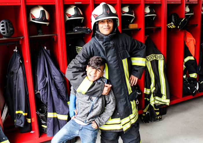 KI generiert: Das Bild zeigt eine Feuerwehrfrau in voller Schutzausrüstung, die einen lächelnden Jungen umarmt, in einer Feuerwache vor roten Schränken mit Feuerwehrhelmen und -kleidung. Der Hauptinhalt des Bildes ist die herzliche Interaktion zwischen der Feuerwehrfrau und dem Jungen in einer Feuerwache.