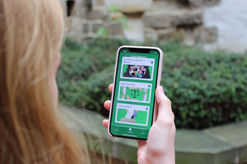 KI generiert: Das Bild zeigt eine Person von hinten, die ein Smartphone mit einer grünen App-Oberfläche hält, auf der verschiedene Bilder und Texte zu sehen sind. Der Hauptinhalt des Bildes ist die Nutzung einer mobilen Anwendung, möglicherweise für Bildungs- oder Informationszwecke.