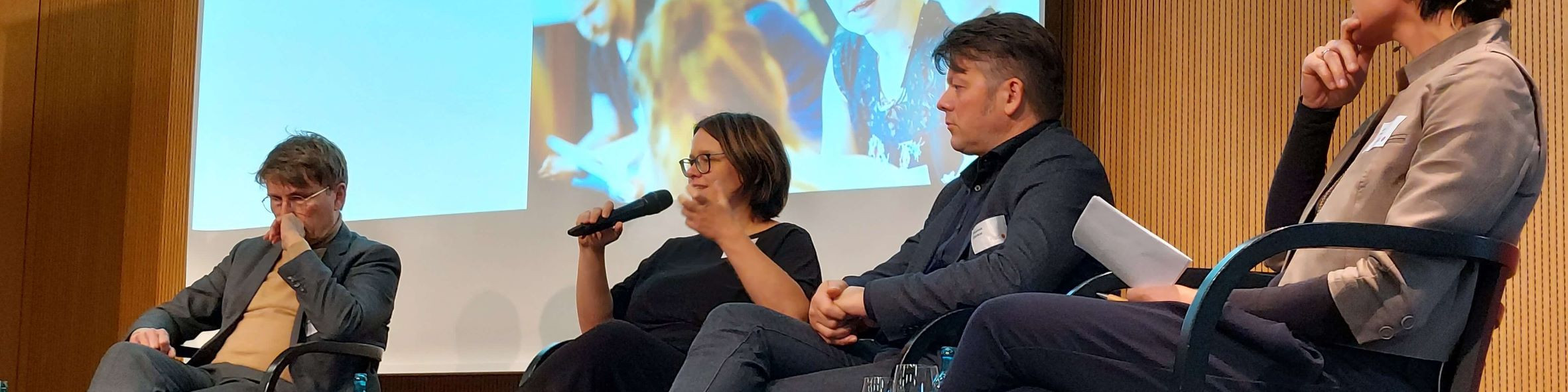 KI generiert: Das Bild zeigt eine Podiumsdiskussion mit vier Personen, die auf der Bühne sitzen und sich unterhalten, während eine Präsentation oder ein Video auf einer Leinwand im Hintergrund läuft. Der Hauptinhalt des Bildes ist die Diskussion oder das Panel auf der Bühne.