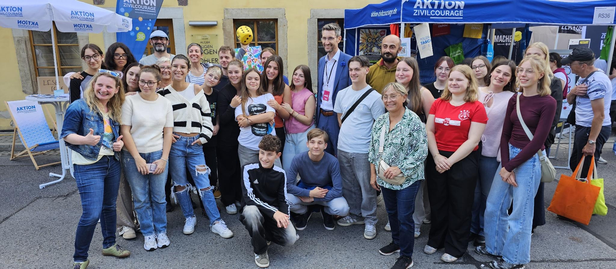 KI generiert: Das Bild zeigt eine Gruppe von Menschen, die vor einem Gebäude mit einem "AKTION ZIVILCOURAGE" Banner posieren. Es handelt sich vermutlich um eine Veranstaltung oder ein Treffen im Rahmen einer zivilgesellschaftlichen Initiative.
