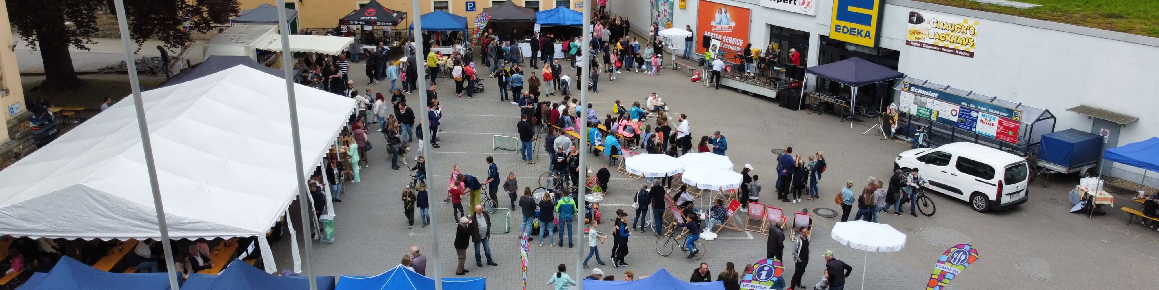 KI generiert: Das Bild zeigt eine belebte Outdoor-Veranstaltung mit vielen Menschen, die zwischen Ständen, Zelten und einem Gebäude mit Werbebannern umherlaufen. Im Vordergrund sind weiße Sonnenschirme und ein Fahrrad zu sehen, während im Hintergrund weitere Besucher und Aussteller zu erkennen sind.