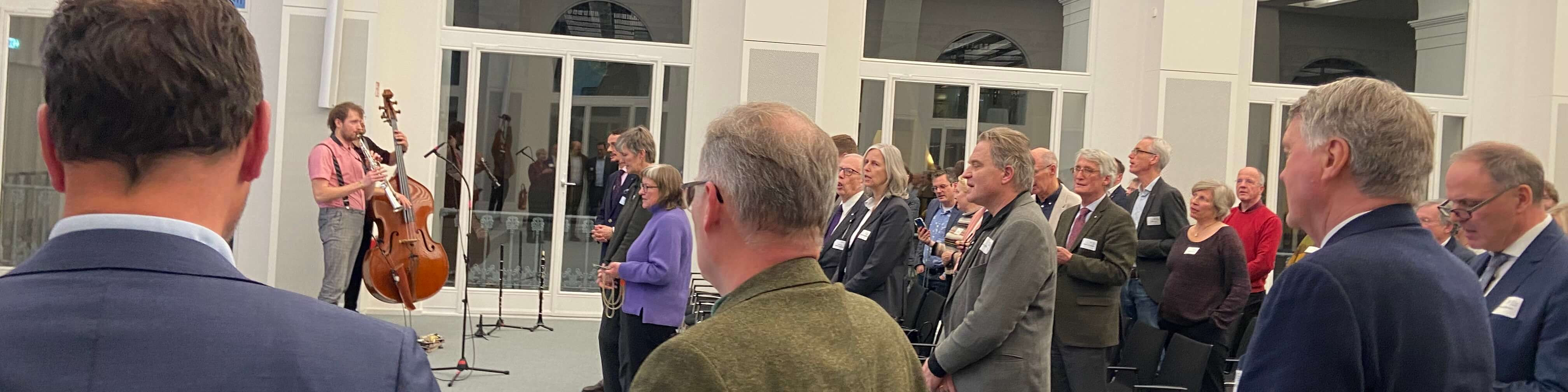 KI generiert: Das Bild zeigt eine Gruppe von Menschen, die einer Musikdarbietung mit einem Cellisten und einem Kontrabassisten in einem hellen, modernen Innenraum zuhören. Die Zuhörer stehen und scheinen aufmerksam dem Konzert zu folgen.
