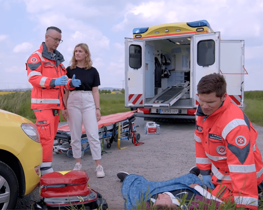 KI generiert: Das Bild zeigt eine Notfallsituation, bei der zwei Rettungssanitäter in orangefarbener Schutzkleidung und eine Frau neben einem Unfallfahrzeug und einem offenen Rettungswagen stehen. Der Hauptinhalt des Bildes ist die Darstellung einer Rettungsaktion nach einem Verkehrsunfall.