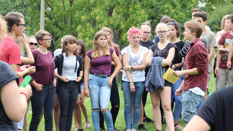 KI generiert: Das Bild zeigt eine Gruppe junger Menschen, die im Freien stehen und sich unterhalten. Der Hauptinhalt ist eine gesellige Zusammenkunft in einem parkähnlichen Umfeld.