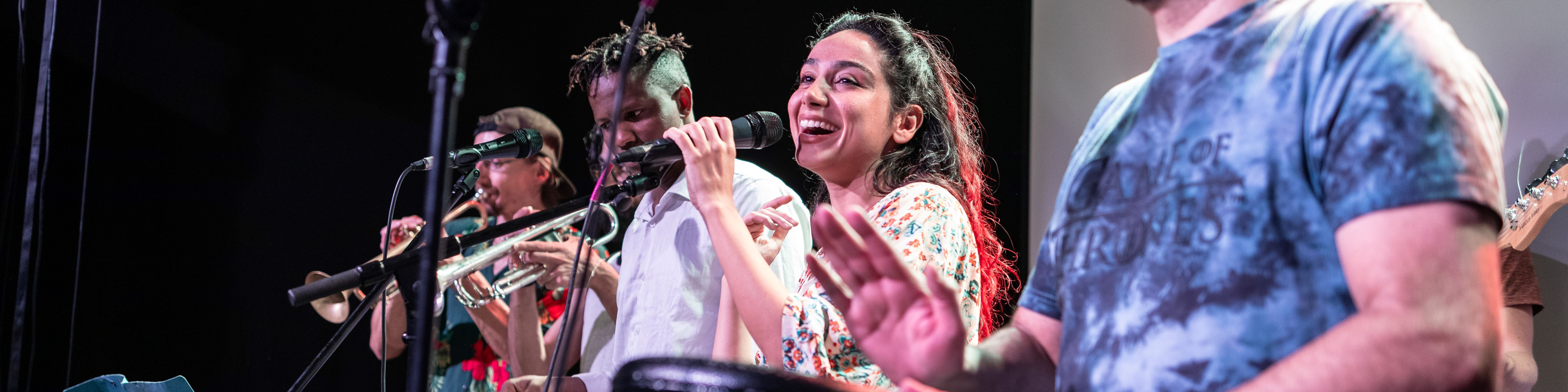 KI generiert: Das Bild zeigt eine lebhafte Band, die auf der Bühne spielt, mit einer Sängerin im Vordergrund, die in ein Mikrofon singt, während andere Musiker verschiedene Instrumente wie Trompete und Schlagzeug spielen. Der Hauptinhalt des Bildes ist die Darstellung einer energiegeladenen Live-Musikperformance.