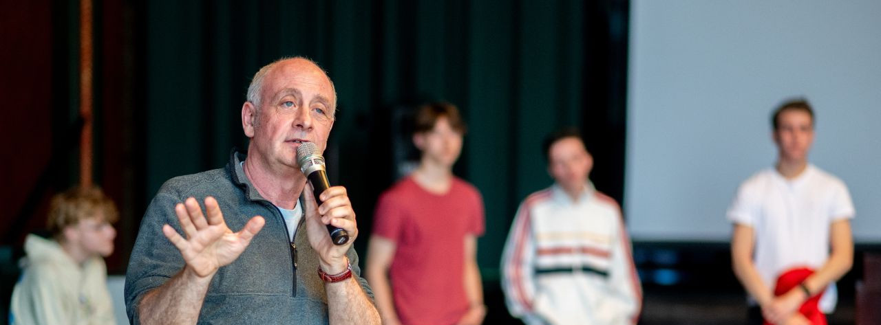 KI generiert: Das Bild zeigt einen Mann, der auf einer Bühne steht und in ein Mikrofon spricht, während er eine Handbewegung macht. Im Hintergrund sind mehrere Personen, die ihm zuhören, sowie eine Leinwand zu sehen.