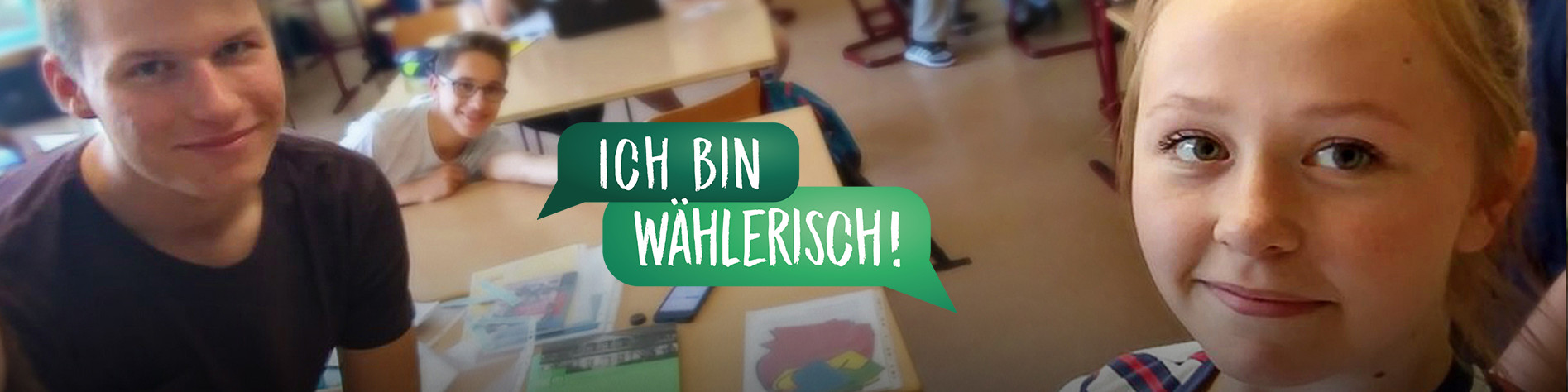 KI generiert: Das Bild zeigt zwei Personen in einem Klassenzimmer, wobei eine Person an einem Tisch sitzt und die andere Person in der Nähe steht. Im Vordergrund steht der Text "ICH BIN WÄHLERISCH!" in einem grünen Sprechblasen-Design.
