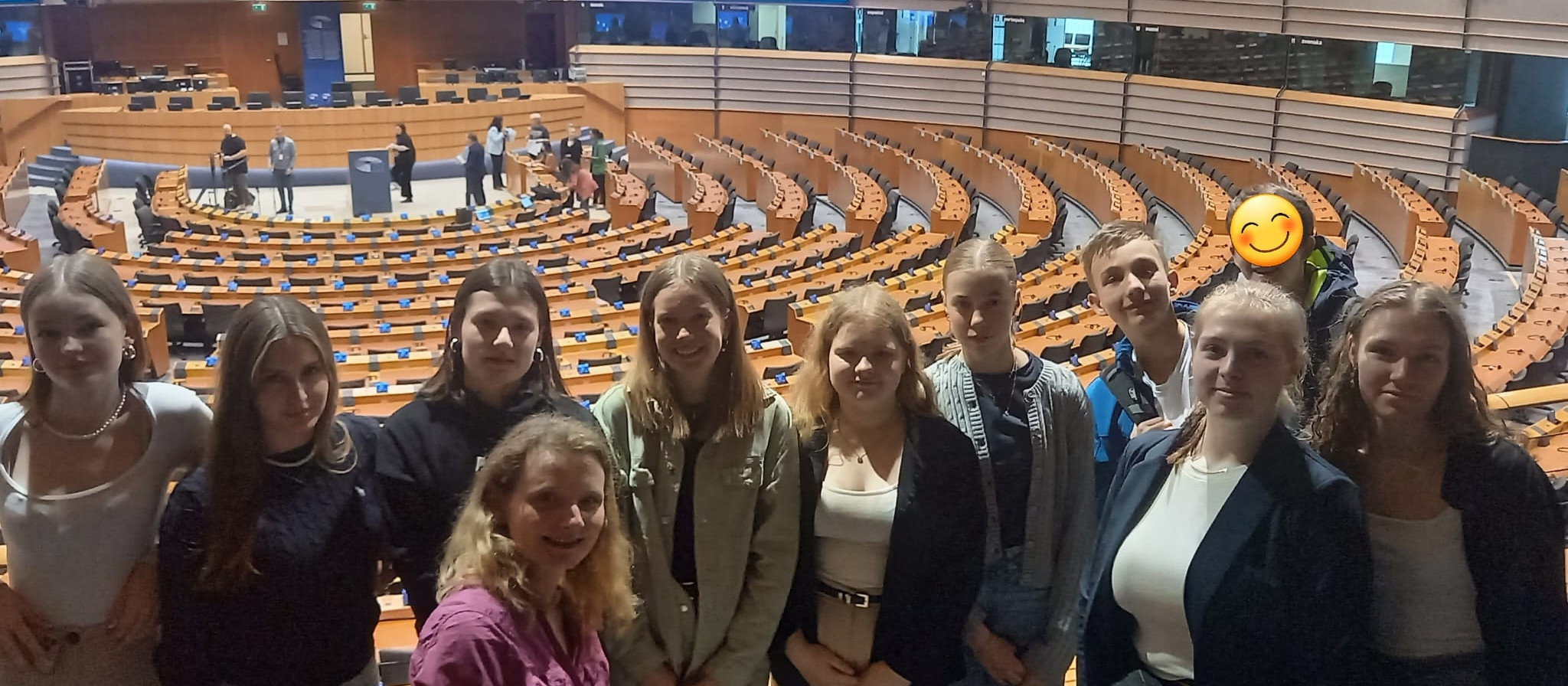 KI generiert: Das Bild zeigt eine Gruppe von Personen, die in einem großen, halbrunden Konferenzsaal mit vielen leeren Sitzen posieren. Der Hauptinhalt des Bildes ist die Gruppe von Menschen, die vor dem Hintergrund eines offiziellen oder politischen Versammlungsraums steht.