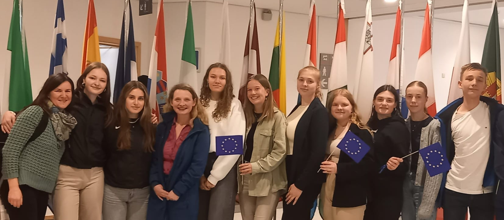 KI generiert: Das Bild zeigt eine Gruppe von Personen, die vor einer Reihe von Flaggen verschiedener Länder posieren. Der Hauptinhalt des Bildes ist die Darstellung einer internationalen oder europäischen Zusammenkunft, wobei einige Personen die Flagge der Europäischen Union halten.