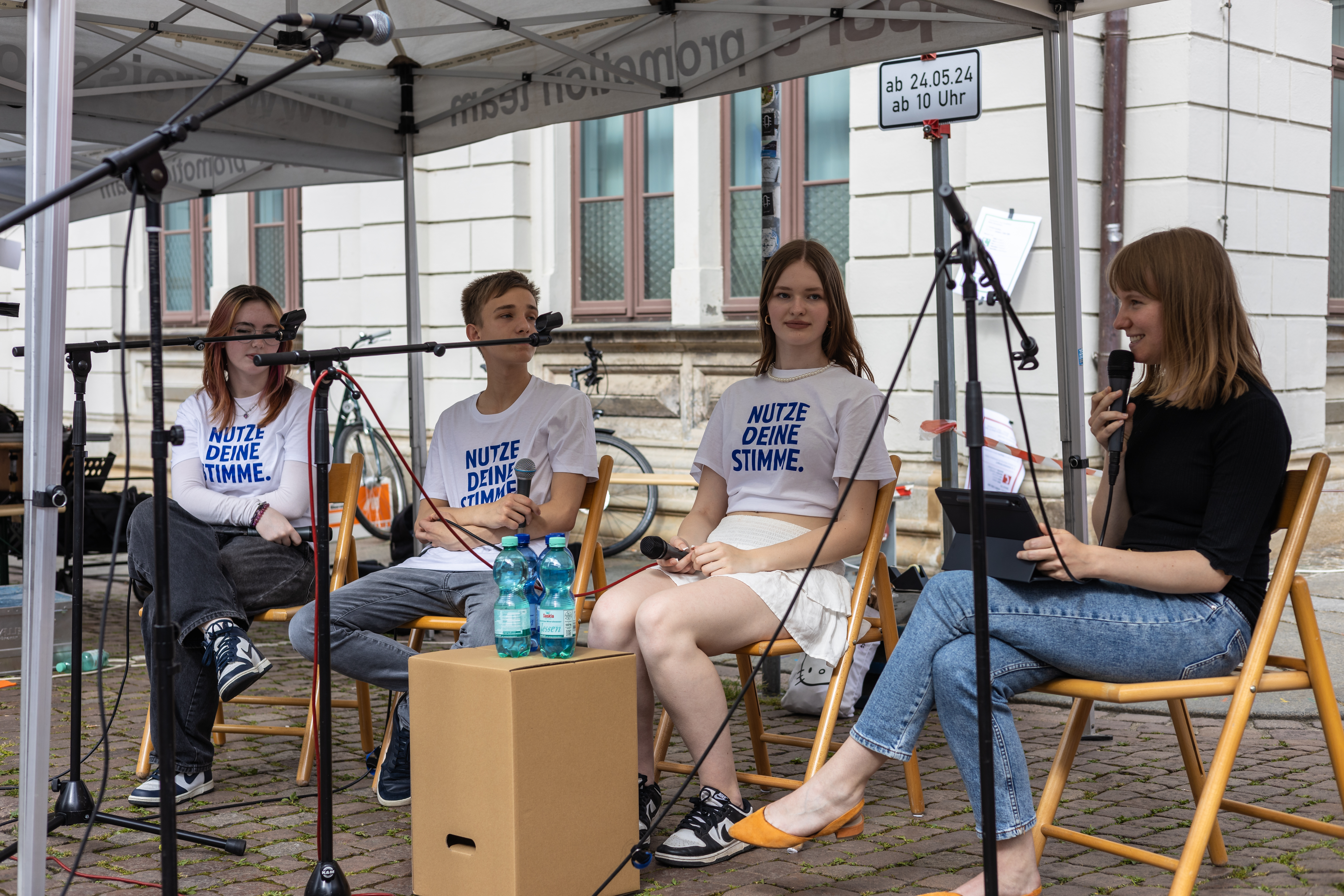 KI generiert: Das Bild zeigt vier Personen, die unter einem Zelt auf Stühlen sitzen und eine Diskussion oder ein Interview führen, wobei drei von ihnen T-Shirts mit der Aufschrift "NUTZE DEINE STIMME" tragen. Im Vordergrund sind Mikrofone und ein Wasserflasche auf einem kleinen Tisch zu sehen, was auf eine öffentliche Veranstaltung oder einen Podcast hinweist.