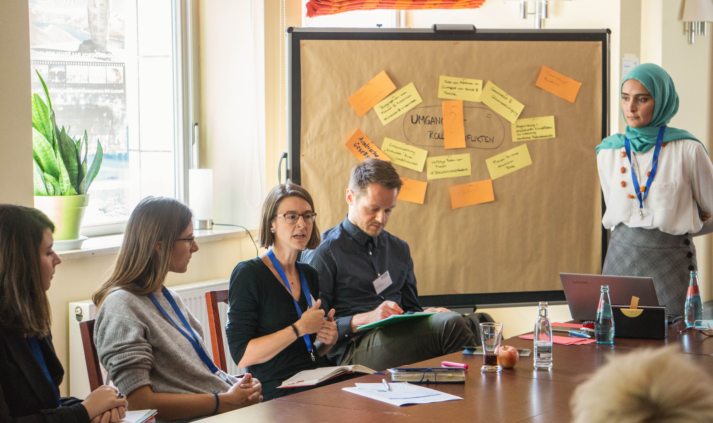 KI generiert: Das Bild zeigt eine Gruppe von Personen in einem Besprechungsraum, die an einem Tisch sitzen und an einer Diskussion oder einem Workshop teilnehmen. Eine Person steht und präsentiert Ideen, die auf einem Flipchart mit Post-its notiert sind.