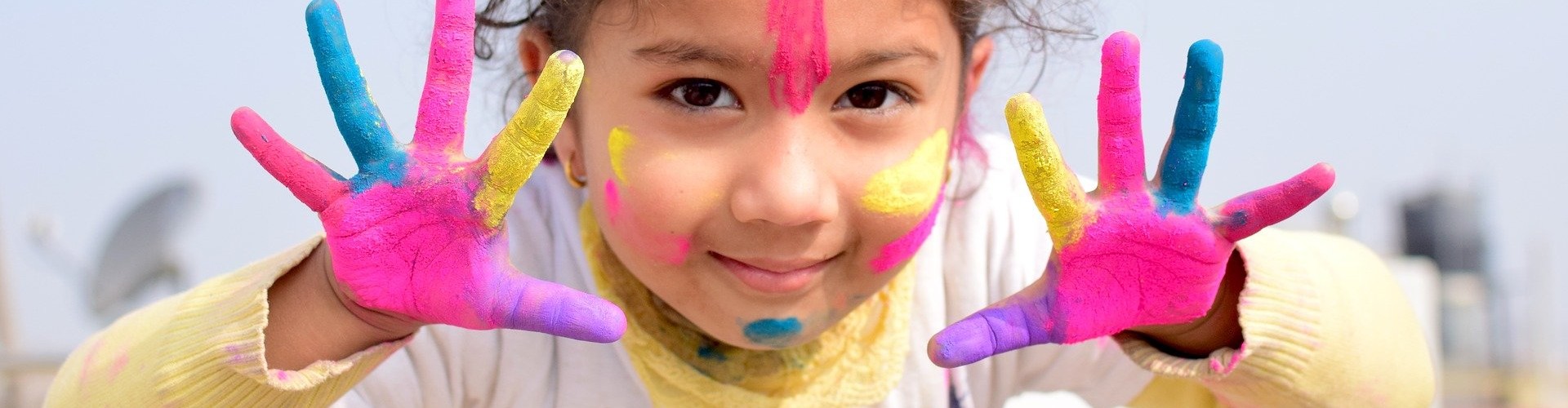 KI generiert: Ein fröhliches Kind zeigt seine bunt bemalten Hände und hat auch Farbe im Gesicht. Es strahlt Freude und Verspieltheit aus, wahrscheinlich während eines farbenfrohen Festes.