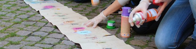 KI generiert: Das Bild zeigt eine Person, die auf einem mit Papier bedeckten Boden kniet und mit mehreren Spraydosen arbeitet. Der Hauptinhalt des Bildes ist das Graffiti-Sprühen oder die Erstellung von Street Art.