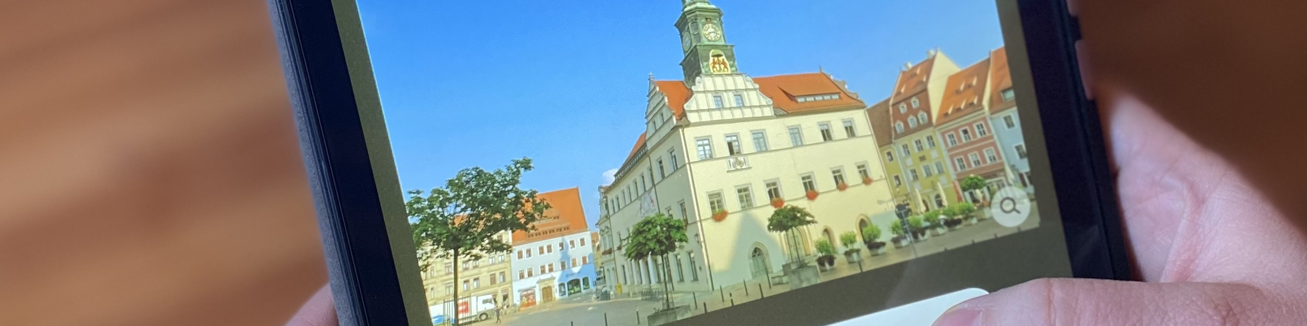 KI generiert: Das Bild zeigt eine Person, die ein Smartphone hält, auf dem ein Foto einer malerischen Stadt mit historischen Gebäuden und einem markanten Rathaus mit Uhrturm zu sehen ist. Der Hauptinhalt des Bildes ist die auf dem Smartphone angezeigte Stadtansicht.