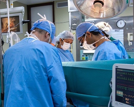 KI generiert: Das Bild zeigt ein medizinisches Team, das sich in einem Operationssaal auf eine Operation vorbereitet. Im Vordergrund sind Ärzte und Pflegekräfte in OP-Kleidung zu sehen, während im Hintergrund medizinische Geräte und ein Patient auf dem OP-Tisch liegen.