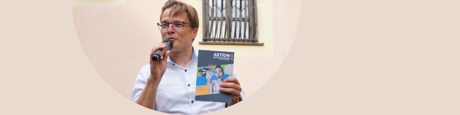 KI generiert: Das Bild zeigt eine Person, die ein Mikrofon in der Hand hält und ein Heft oder eine Broschüre mit dem Titel "AKTION" präsentiert. Im Hintergrund ist ein Fenster mit Gittern zu sehen.