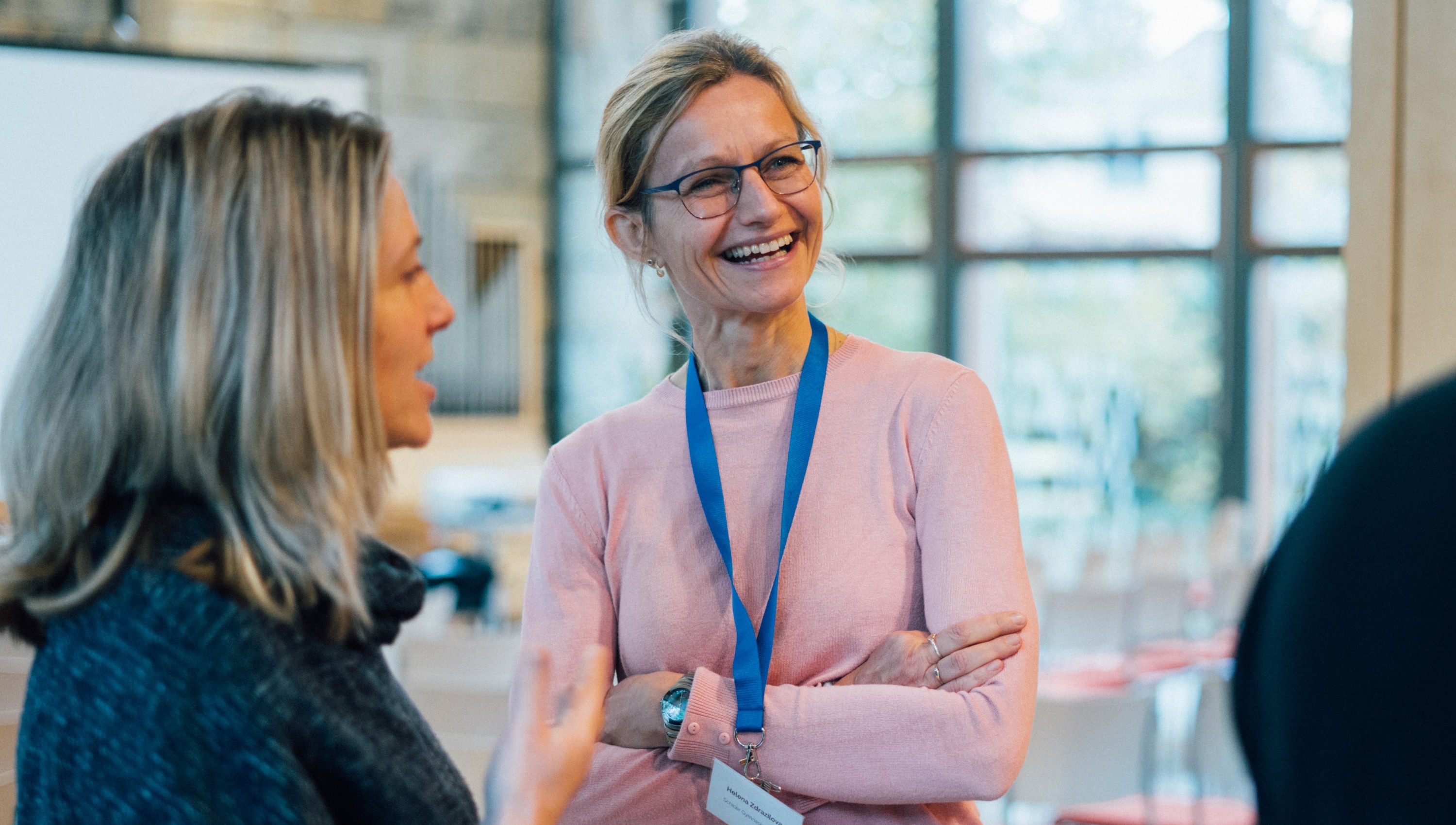 KI generiert: Das Bild zeigt zwei Frauen in einem hellen, modernen Innenraum, die sich angeregt unterhalten. Eine der Frauen trägt ein rosafarbenes Oberteil und eine blaue Namensschilder-Schnur um den Hals.