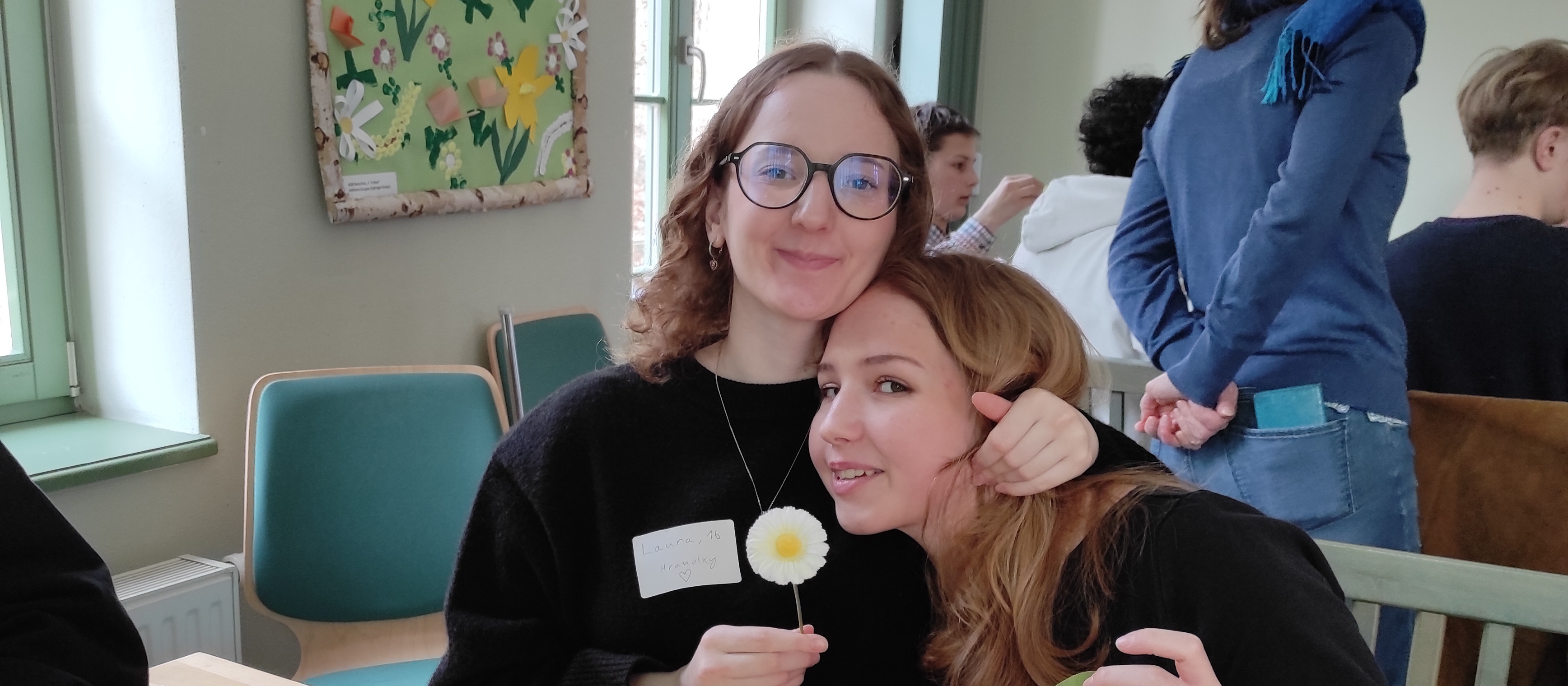 KI generiert: Das Bild zeigt zwei lächelnde Frauen, die sich umarmen, wobei eine von ihnen eine kleine Blume in der Hand hält. Im Hintergrund sind weitere Personen in einem Raum mit Fenstern und einer Wanddekoration zu sehen.