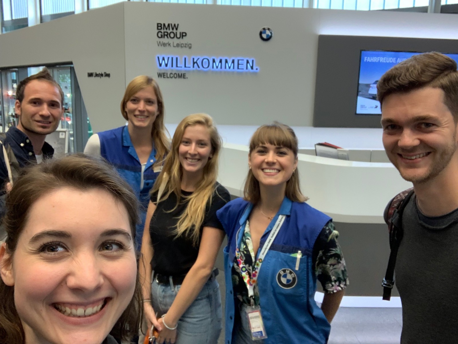 KI generiert: Das Bild zeigt eine Gruppe von sechs Personen, die sich vor einem BMW Group Empfangsbereich mit einem Willkommensschild auf Deutsch und Englisch fotografieren lassen. Die Personen wirken fröhlich und einige tragen Kleidung mit BMW-Logos.