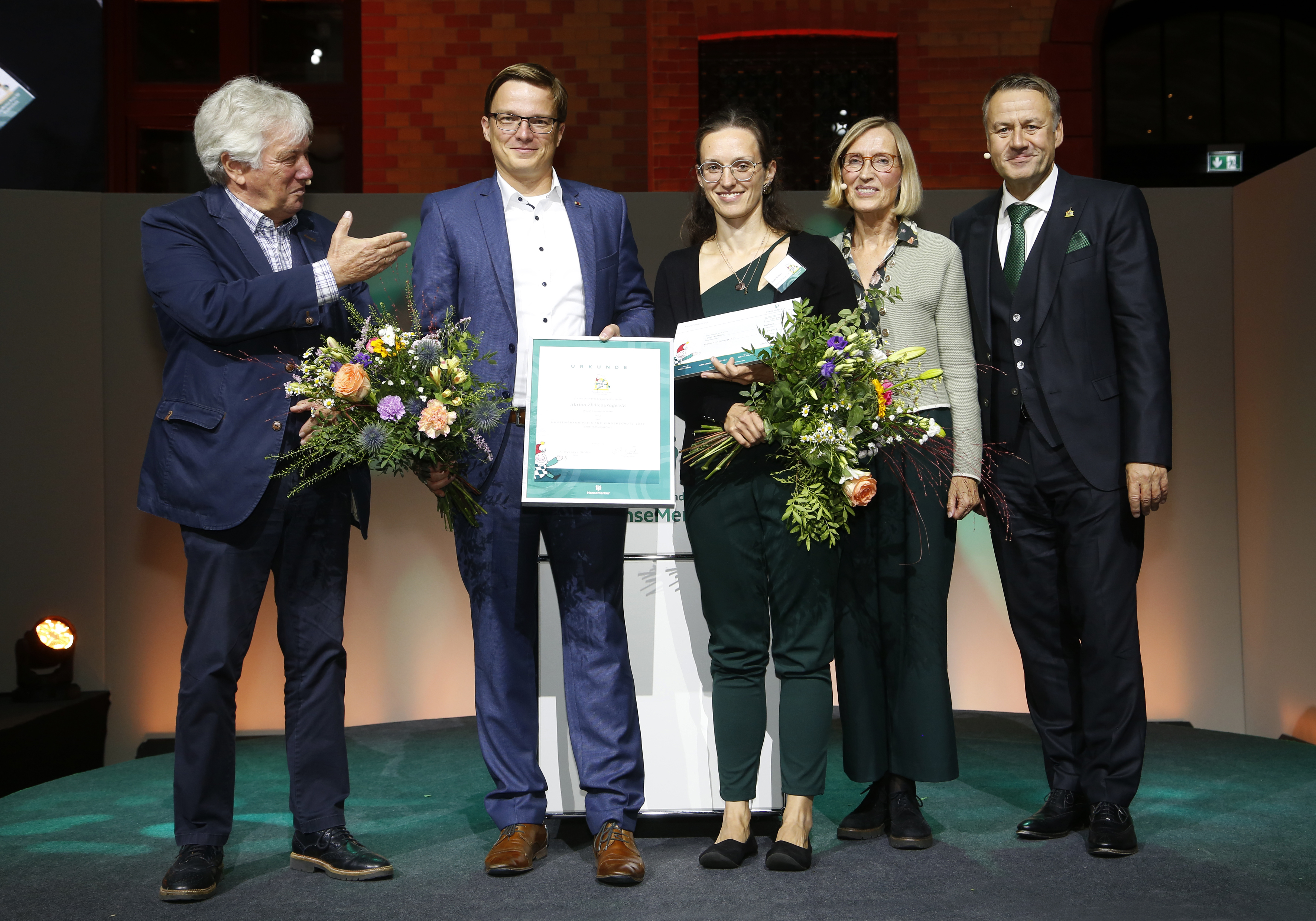 KI generiert: Das Bild zeigt eine Gruppe von fünf Personen auf einer Bühne, wobei zwei Frauen in der Mitte jeweils Blumensträuße und Urkunden in den Händen halten. Es handelt sich vermutlich um eine Preisverleihung oder eine offizielle Ehrung.