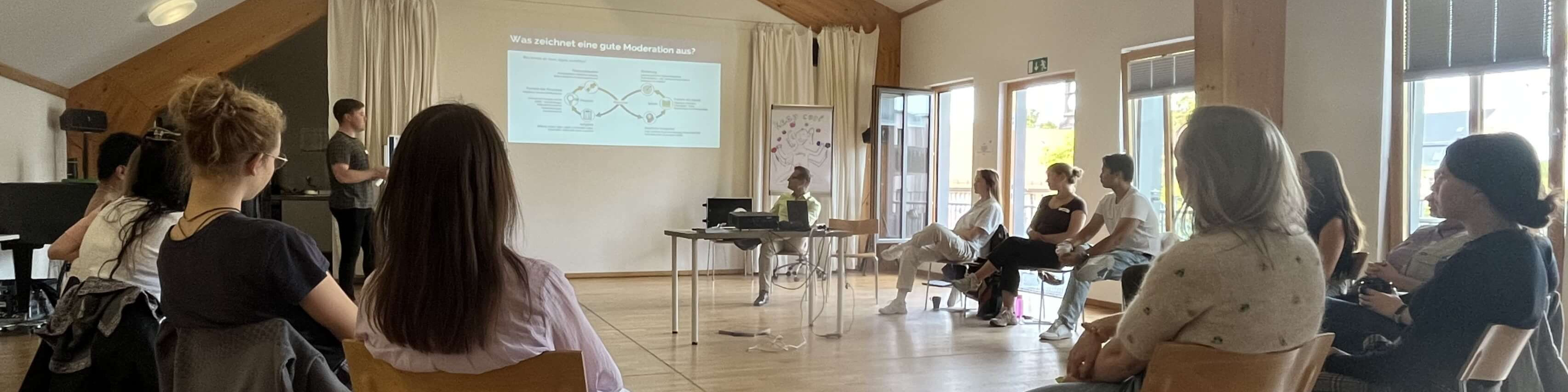 KI generiert: Das Bild zeigt eine Gruppe von Personen in einem Seminarraum, wobei eine Person vorne steht und eine Präsentation hält, während die anderen zuhören. Der Hauptinhalt des Bildes ist eine Bildungs- oder Schulungsveranstaltung.