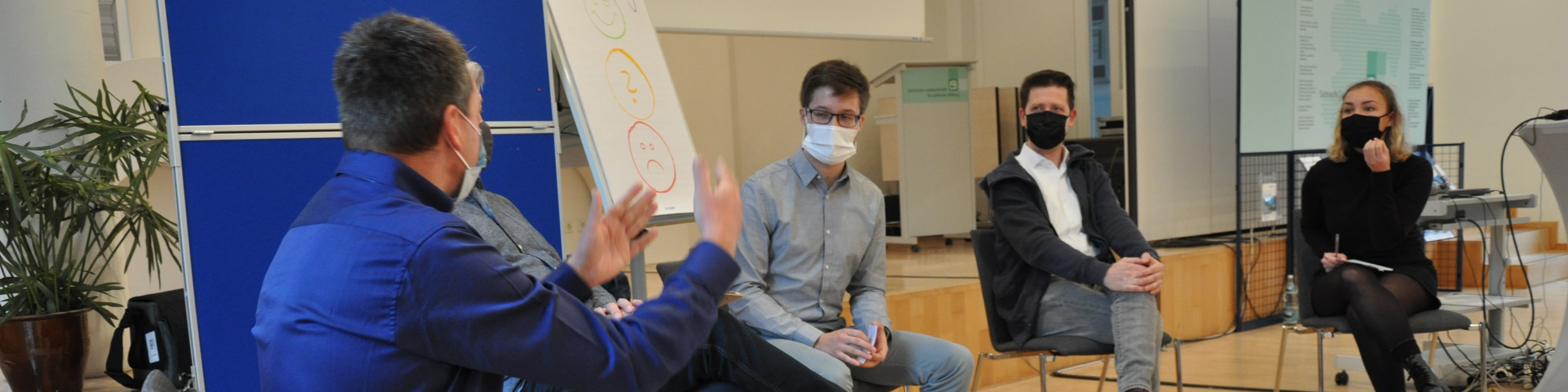 KI generiert: Das Bild zeigt eine Gruppe von Personen in einem Seminarraum, wobei eine Person in einem blauen Jackett steht und eine Präsentation hält, während die anderen Teilnehmer, die alle Masken tragen, zuhören. Der Hauptinhalt des Bildes ist eine interaktive Präsentation oder Diskussion in einem geschäftlichen oder akademischen Umfeld.