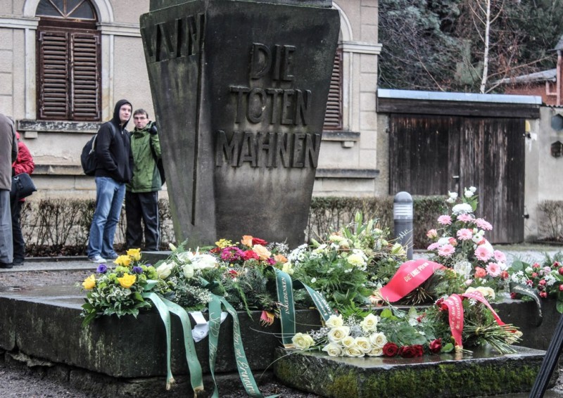 KI generiert: Das Bild zeigt ein Denkmal mit der Inschrift "DIE TOTEN MAHNEN" (Die Toten ermahnen), das mit zahlreichen Blumen und Kränzen geschmückt ist. Im Hintergrund stehen zwei Personen vor einem Gebäude mit geschlossenen Fensterläden.