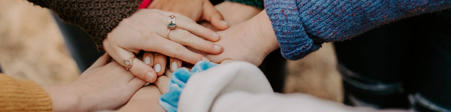 KI generiert: Das Bild zeigt mehrere Hände, die sich in der Mitte übereinanderlegen, was Symbol für Zusammenhalt und Teamgeist steht. Die Personen tragen verschiedene Kleidungsstücke, darunter ein blauer Pullover und ein brauner Mantel.