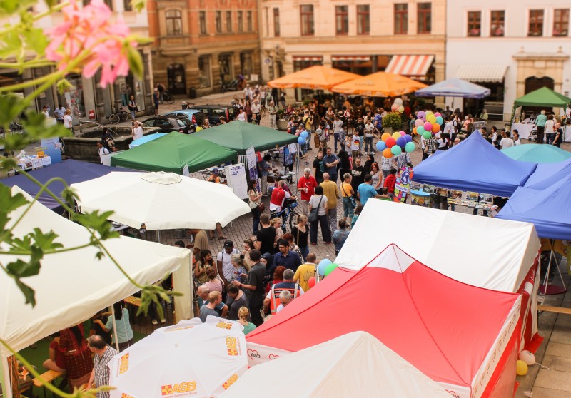 KI generiert: Das Bild zeigt eine belebte Straßenveranstaltung oder ein Fest mit zahlreichen Menschen, die zwischen bunten Zelten und Ständen umherlaufen. Im Vordergrund sind verschiedene farbige Markisen und Luftballons zu sehen, die eine fröhliche und lebendige Atmosphäre vermitteln.