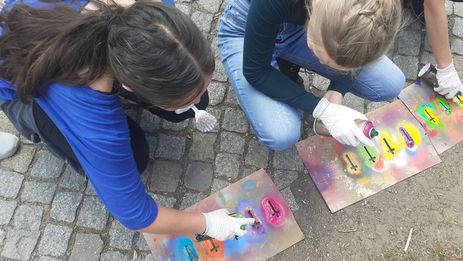 KI generiert: Das Bild zeigt zwei Personen, die auf einem gepflasterten Boden kniend mit Spraydosen und Schablonen Graffiti auf Pappkartons sprühen. Der Hauptinhalt des Bildes ist die kreative Tätigkeit des Graffiti-Sprühens.