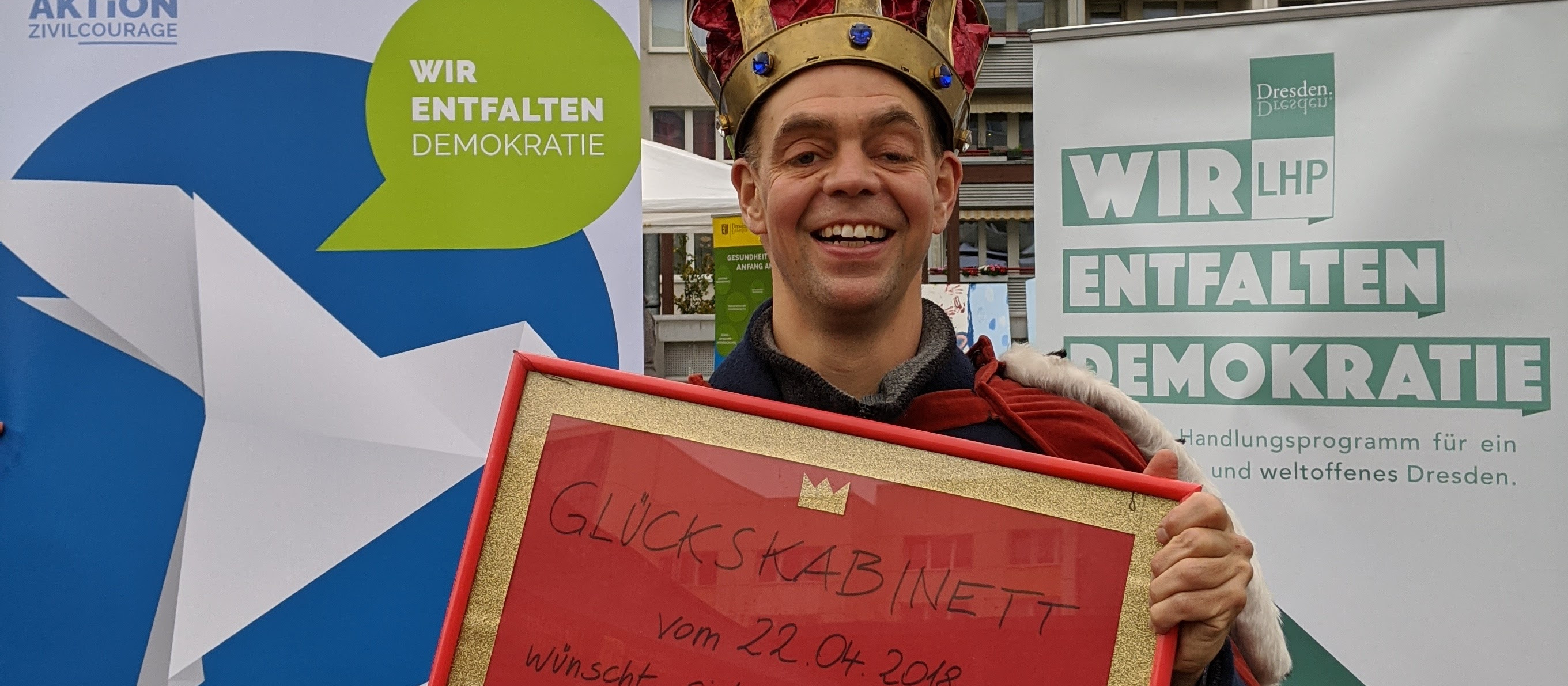 KI generiert: Das Bild zeigt einen lächelnden Mann mit einer Krone, der ein Schild mit der Aufschrift "Glückskabinett" hält, datiert auf den 22.04.2018. Im Hintergrund sind Plakate mit Aufschriften wie "Wir entfalten Demokratie" und Logos von Organisationen zu sehen.