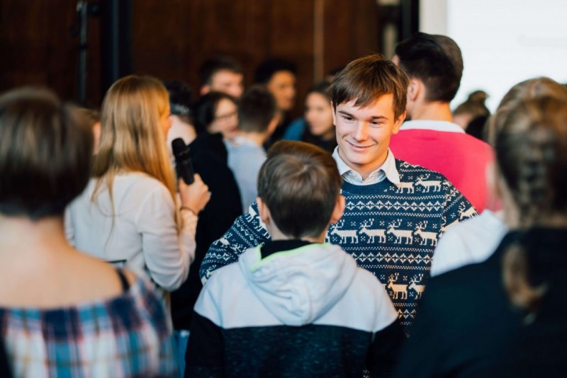 KI generiert: Das Bild zeigt eine Gruppe von Menschen in einem geschäftigen, geselligen Umfeld, wobei ein junger Mann im Vordergrund mit einem reindeer-muster Pullover und ein Kind im Vordergrund zu sehen sind. Der Hauptinhalt des Bildes ist die Interaktion und das Gedränge der Menschenmenge.