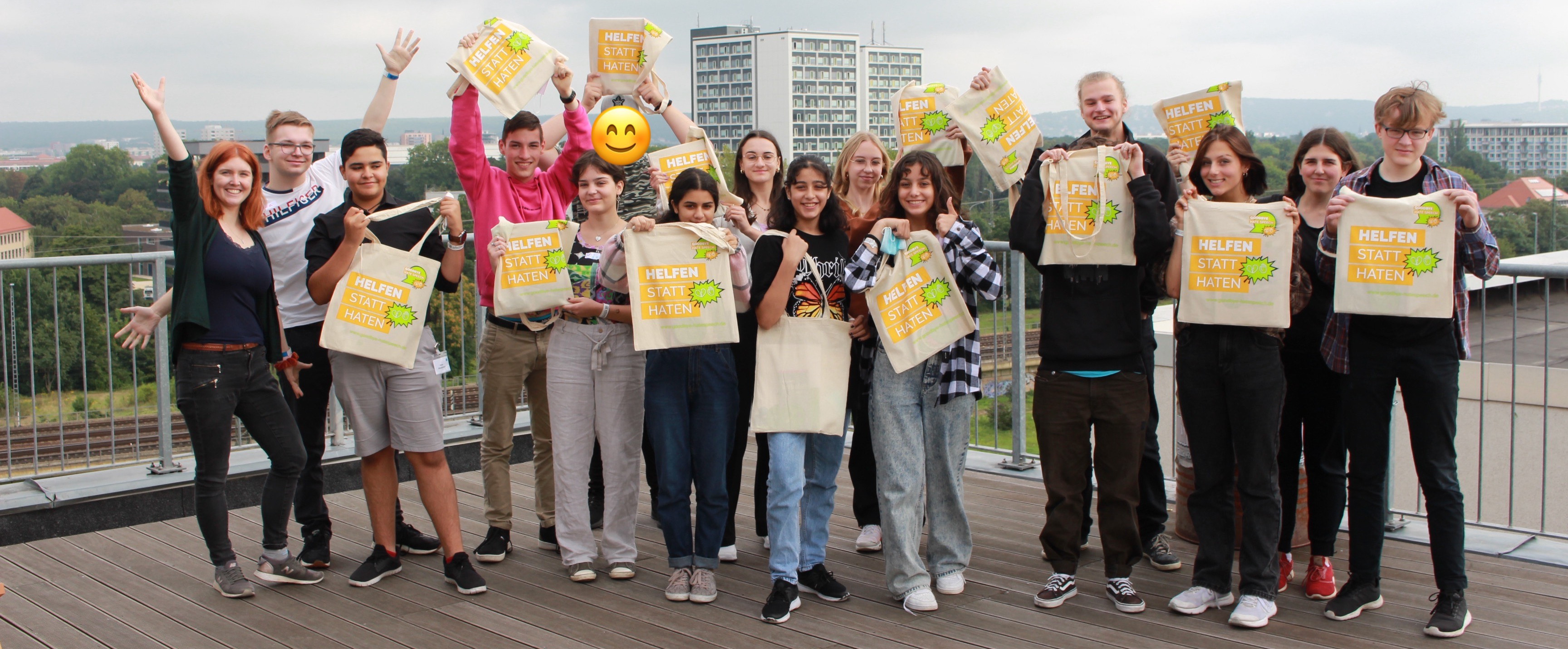 KI generiert: Das Bild zeigt eine Gruppe von Jugendlichen, die auf einer Terrasse stehen und T-Shirts mit der Aufschrift "HELFEN STATT HATEN" hochhalten. Der Hauptinhalt des Bildes ist die gemeinsame Aktion oder Kampagne der Gruppe gegen Hass.