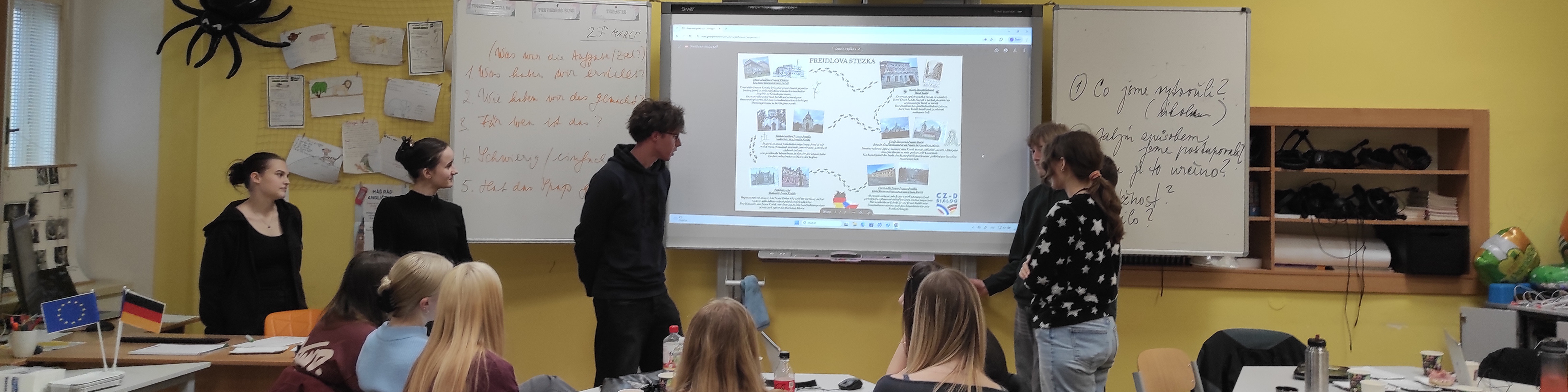 KI generiert: Das Bild zeigt eine Gruppe von Personen in einem Klassenzimmer, wobei eine Person vor einem Whiteboard steht und eine Präsentation hält. Der Hauptinhalt des Bildes ist eine Unterrichts- oder Besprechungssituation, in der eine Präsentation oder Diskussion stattfindet.