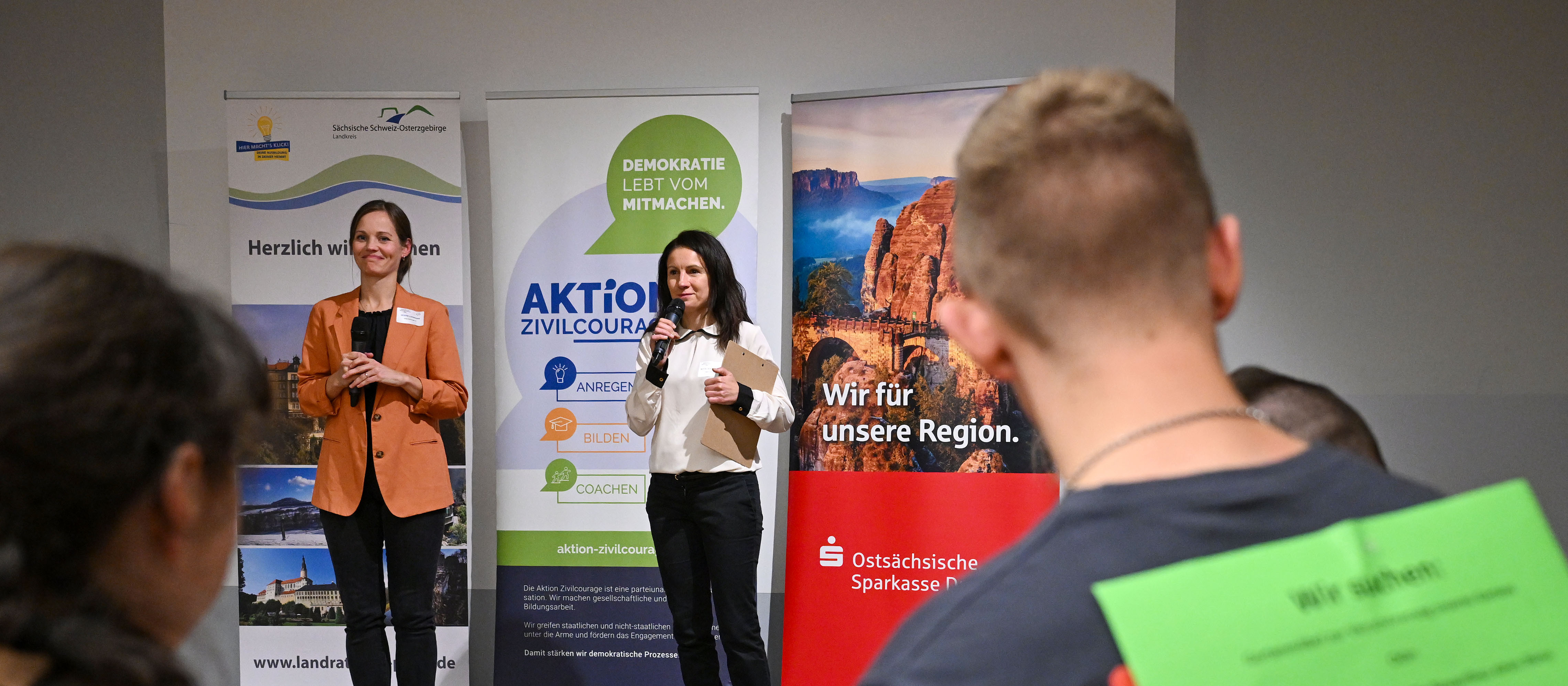 KI generiert: Das Bild zeigt eine Frau, die vor einem Publikum spricht und dabei ein Mikrofon hält. Im Hintergrund sind mehrere Banner mit verschiedenen Logos und Slogans zu sehen, darunter "Demokratie mitmachen" und "Wir für unsere Region".