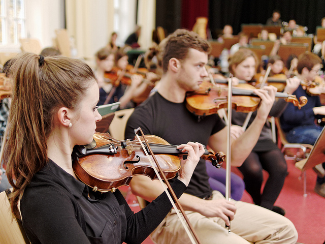 KI generiert: Das Bild zeigt eine Gruppe von Musikern, die in einem Orchester Violinen spielen. Der Hauptinhalt des Bildes ist die Darstellung eines Orchesters bei einer Probe oder Aufführung.