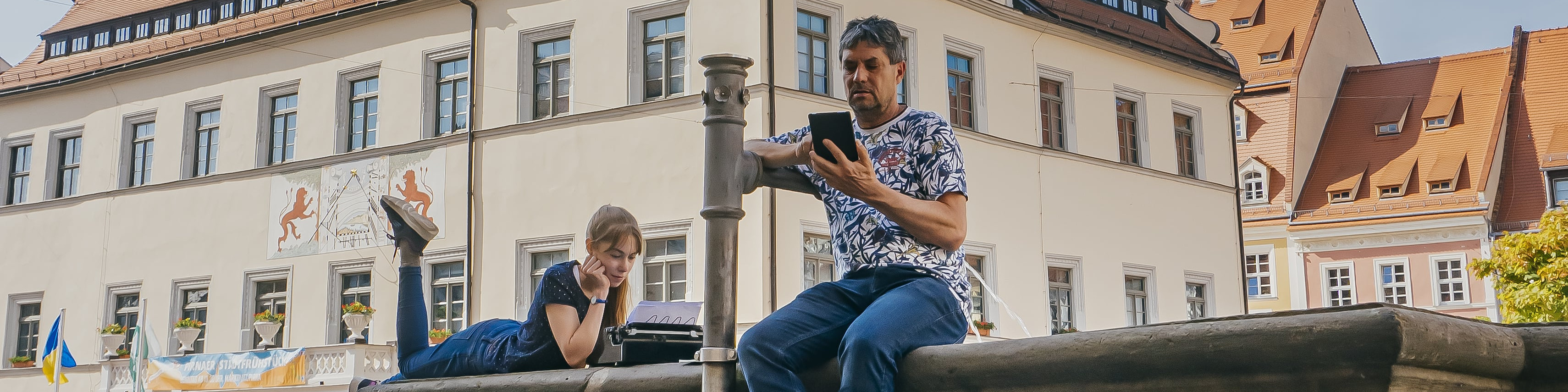 KI generiert: Das Bild zeigt zwei Personen, die auf einem öffentlichen Platz vor historischen Gebäuden sitzen und auf ihre Mobilgeräte schauen. Im Vordergrund ist ein alter Brunnen zu sehen, und im Hintergrund sind traditionelle europäische Häuser mit verzierten Fassaden.
