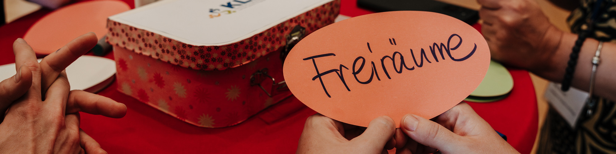 KI generiert: Das Bild zeigt mehrere Hände, die eine orangefarbene Pappform mit der Aufschrift "Freiräume" halten, während im Hintergrund eine rote Geschenkbox mit Polka-Dots zu sehen ist. Der Hauptinhalt des Bildes ist die Betonung von Freiräumen, möglicherweise im Kontext einer Diskussion oder eines Workshops.