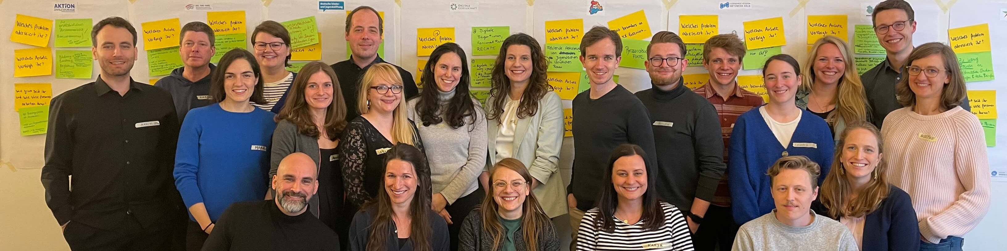 KI generiert: Das Bild zeigt eine Gruppe von Personen, die vor einer Wand mit vielen bunten Notizzetteln posieren. Der Hauptinhalt des Bildes ist eine Team- oder Gruppenfotografie, möglicherweise im Rahmen eines Workshops oder einer Brainstorming-Sitzung.