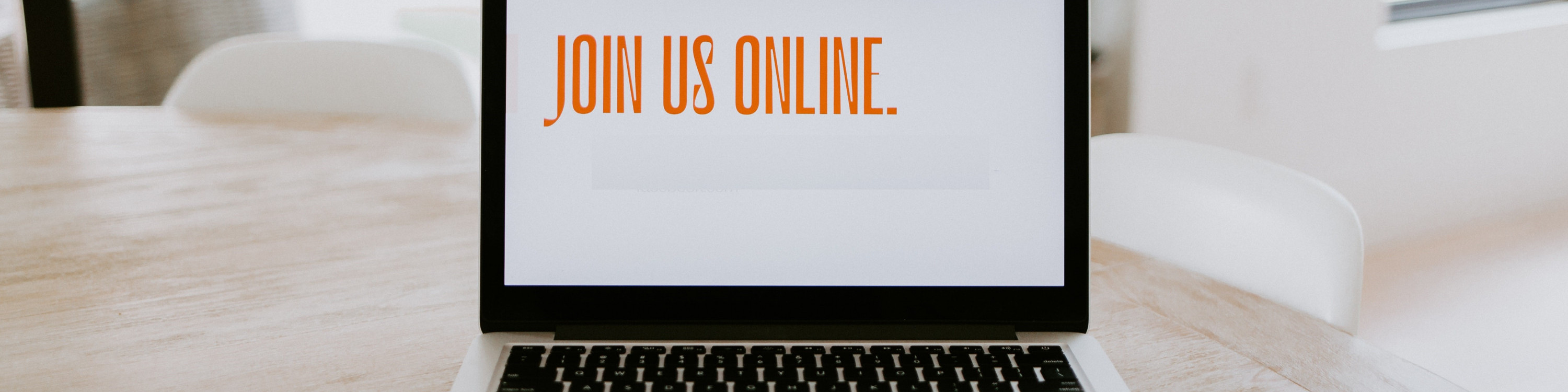 KI generiert: Das Bild zeigt einen Laptop auf einem Holztisch mit der Bildschirmanzeige "JOIN US ONLINE." Im Hintergrund sind unscharf zwei weiße Stühle zu erkennen. Der Hauptinhalt des Bildes ist die Einladung, online teilzunehmen.