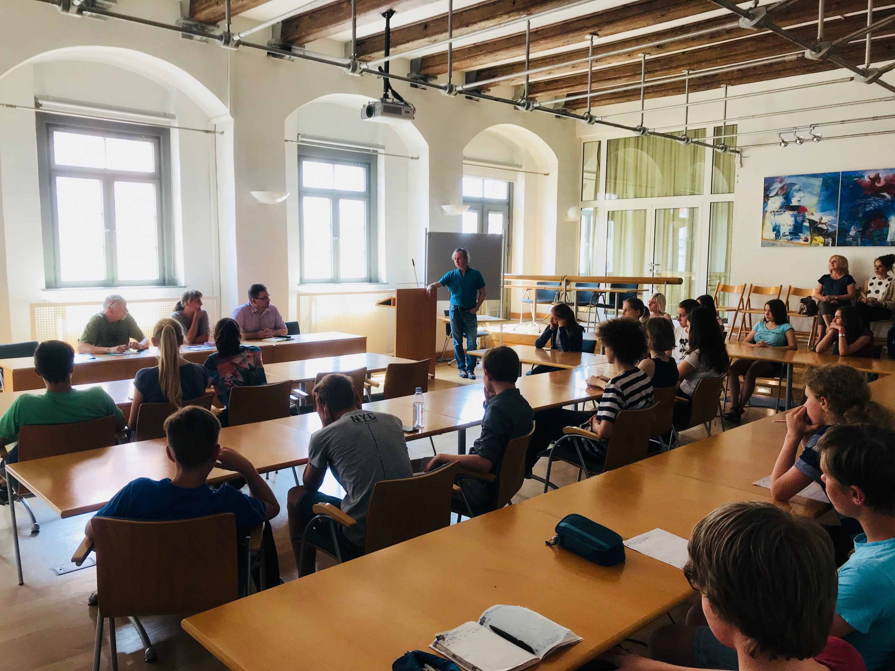 KI generiert: Das Bild zeigt eine Gruppe von Personen in einem hellen Raum mit großen Fenstern und Holzdecken, die an langen Tischen sitzen und einer stehenden Person zuhören, die vorne spricht. Der Hauptinhalt des Bildes ist eine Versammlung oder ein Seminar, bei dem eine Person eine Präsentation oder einen Vortrag hält.