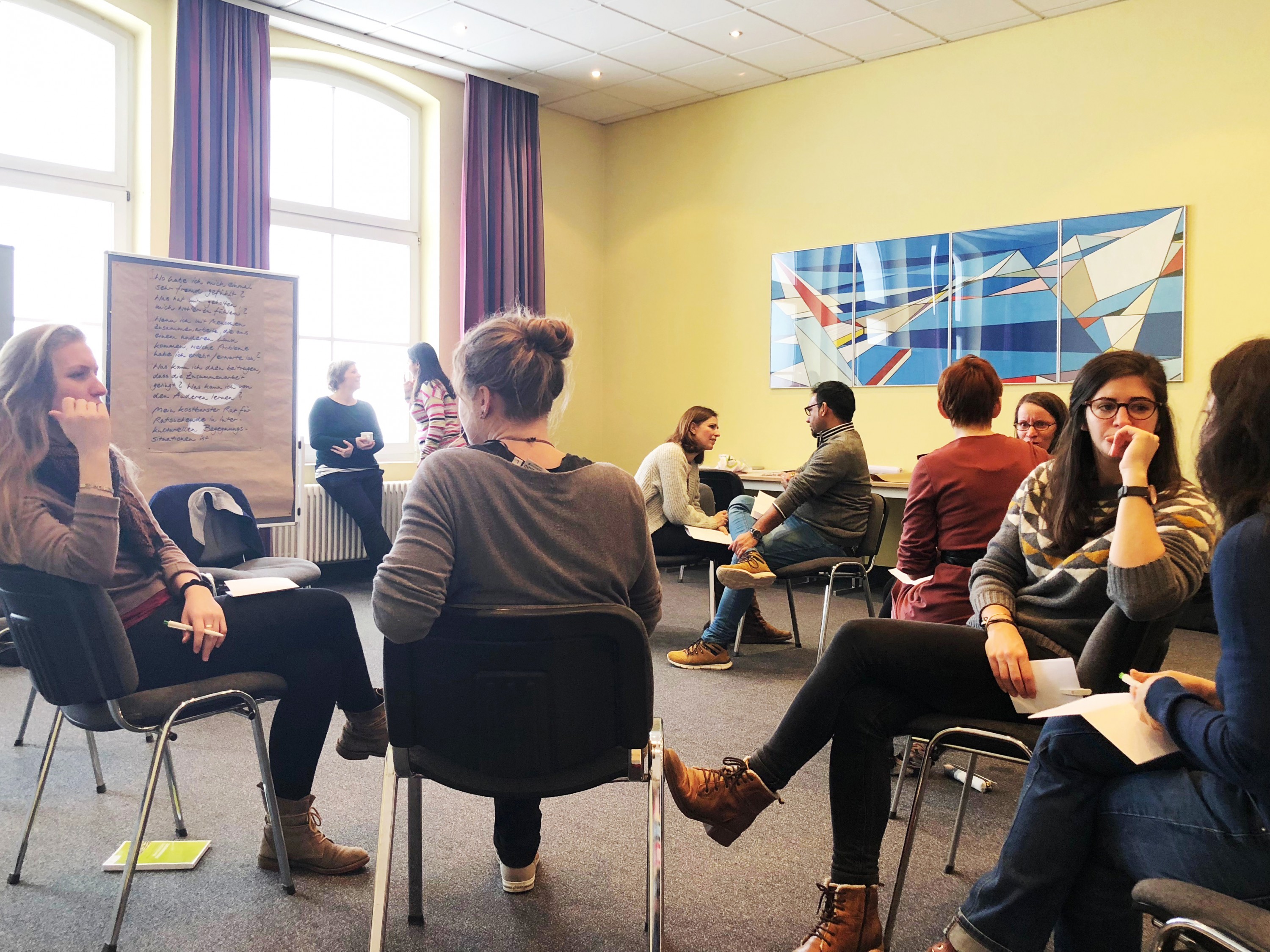 KI generiert: Das Bild zeigt eine Gruppe von Personen, die in einem hellen Raum mit gelben Wänden und großen Fenstern in einem Kreis sitzen und sich unterhalten. Der Hauptinhalt des Bildes ist eine informelle Besprechung oder ein Workshop.