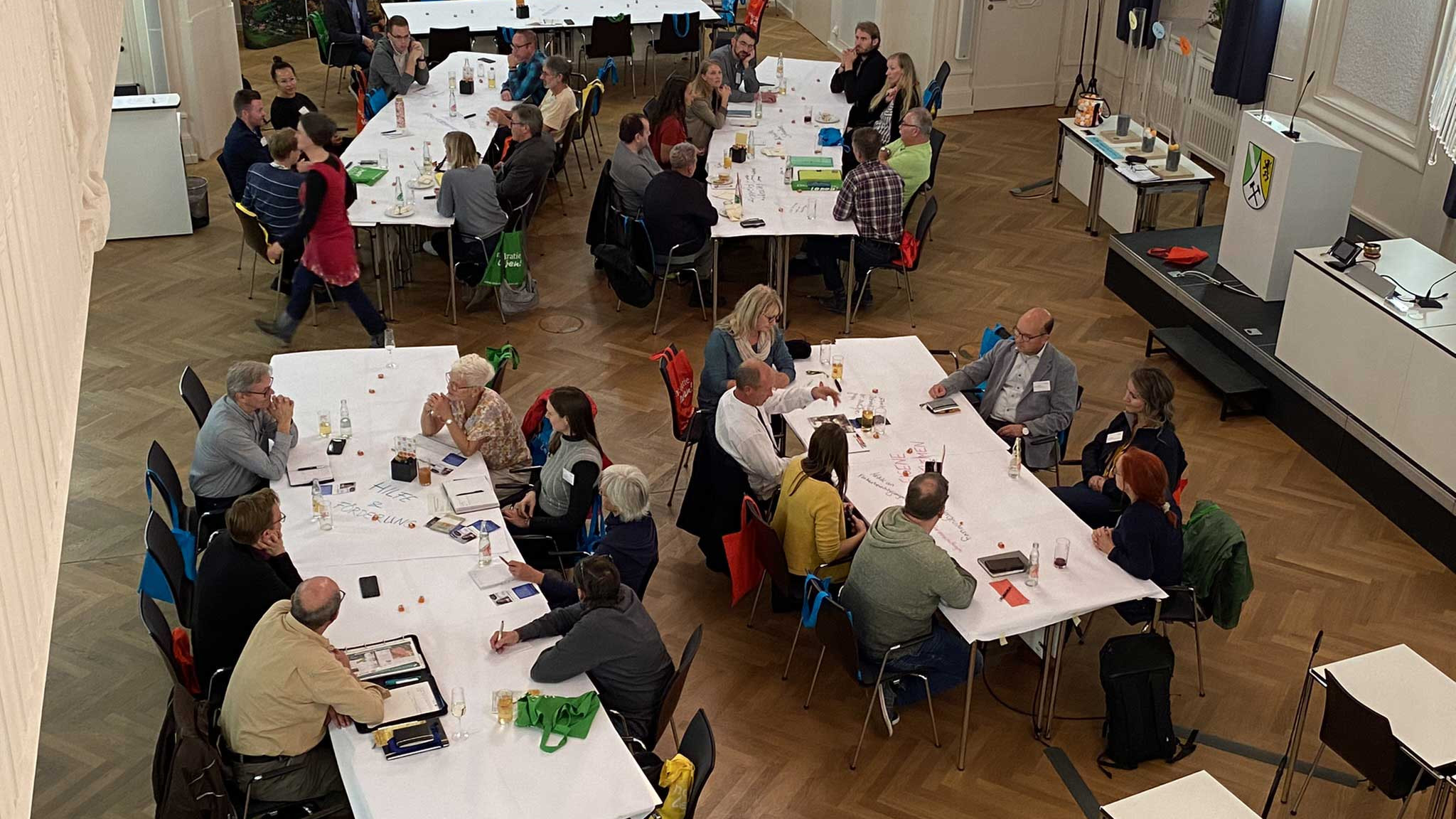 KI generiert: Das Bild zeigt eine Gruppe von Menschen, die in einem großen Raum an langen Tischen sitzen und sich in kleinen Gruppen unterhalten. Der Hauptinhalt des Bildes ist eine Versammlung oder ein Treffen, bei dem die Teilnehmer in einer entspannten Atmosphäre diskutieren und möglicherweise an einer Veranstaltung oder einem Workshop teilnehmen.