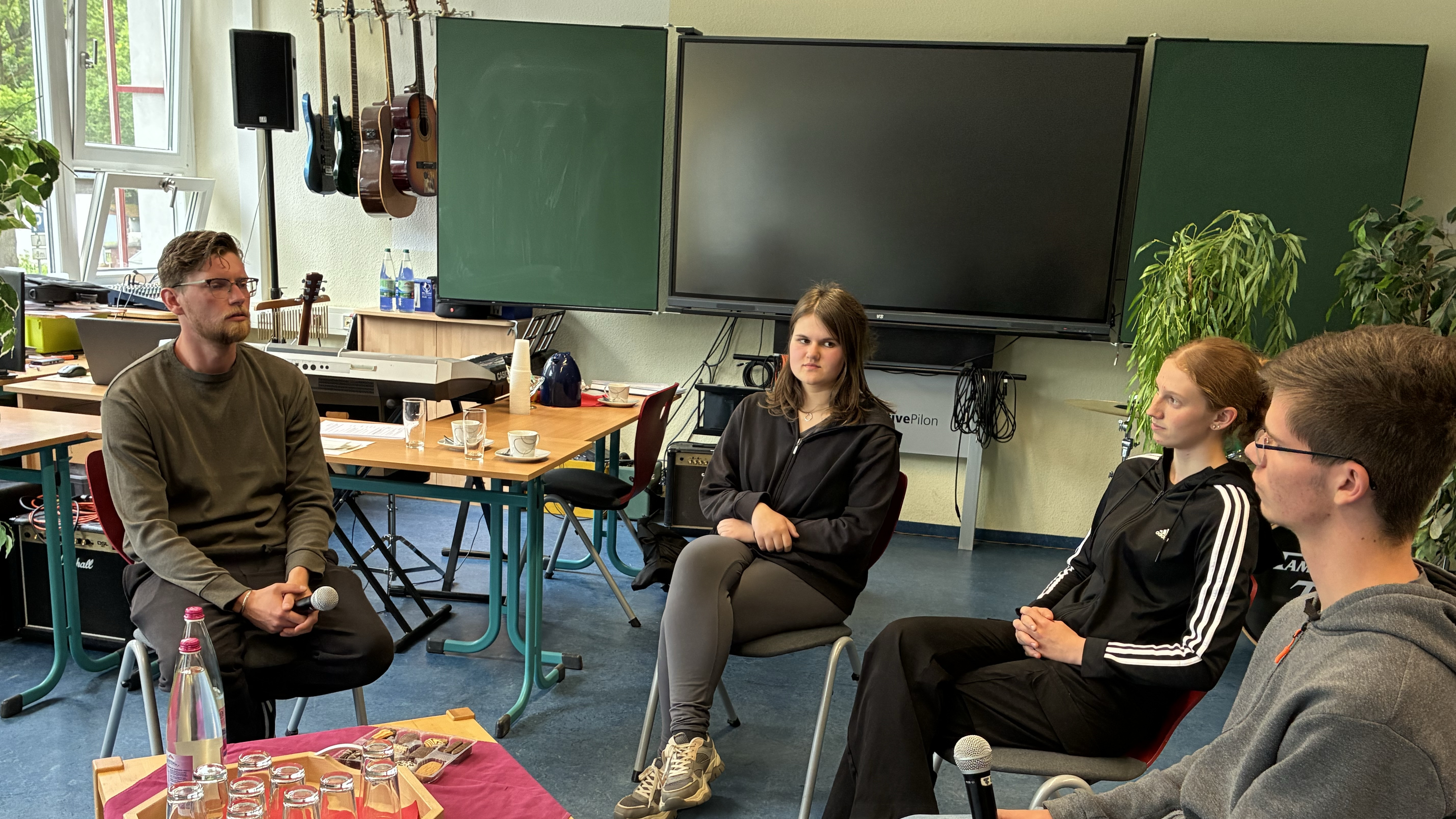 KI generiert: Das Bild zeigt eine Gruppe von vier Personen, die in einem Klassenzimmer an Tischen sitzen und sich unterhalten. Im Hintergrund sind eine Tafel, Musikinstrumente und Pflanzen zu sehen, was auf eine entspannte Diskussion oder ein Meeting in einer schulischen oder kreativen Umgebung hindeutet.
