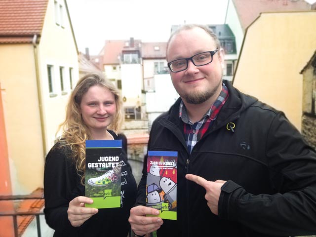 KI generiert: Das Bild zeigt zwei Personen, die jeweils ein Buch in den Händen halten und in die Kamera lächeln. Die Bücher haben bunte Cover mit Titeln, die auf Deutsch geschrieben sind.