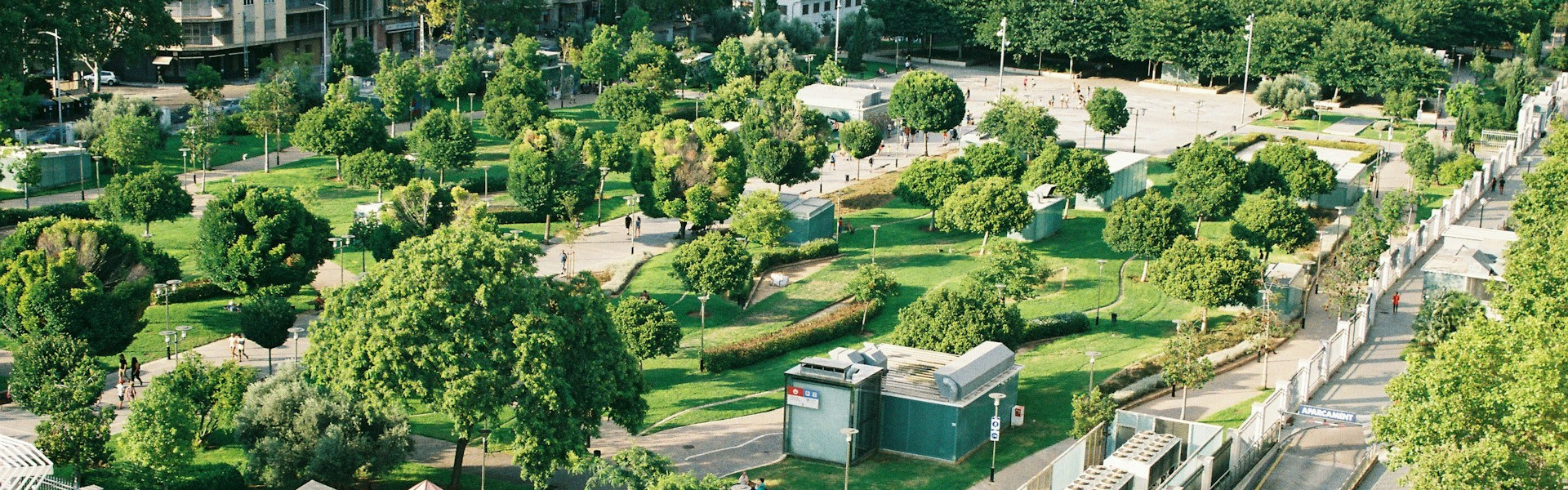 KI generiert: Das Bild zeigt einen weitläufigen, grünen Park mit vielen Bäumen, Spazierwegen und einigen Gebäuden am Rand. Der Hauptinhalt des Bildes ist die idyllische Parklandschaft.