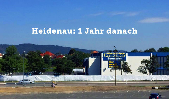 KI generiert: Das Bild zeigt eine Baustelle mit einem großen, abgedeckten Bereich und im Hintergrund ein Gebäude mit einem Praktiker Baumarkt. Über dem Bild steht der Text "Heidenau: 1 Jahr danach".