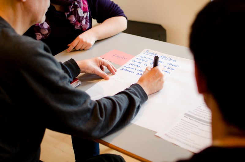 KI generiert: Das Bild zeigt eine Gruppe von Personen, die an einem Tisch sitzen und gemeinsam an einem Projekt arbeiten. Eine Person schreibt mit einem Stift auf ein großes Blatt Papier, während andere zuschauen und mitwirken.