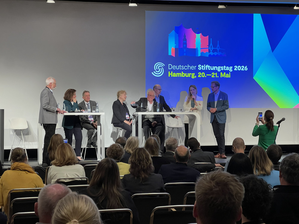 KI generiert: Das Bild zeigt eine Podiumsdiskussion mit mehreren Teilnehmern auf einer Bühne, die vor einem Publikum stattfindet. Im Hintergrund sind eine Leinwand mit den Namen und Titeln der Diskutanten sowie das Logo des Deutschen Fundraising Verbands zu sehen.