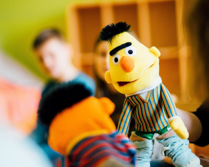 KI generiert: Das Bild zeigt eine Nahaufnahme einer Puppe, die Bert aus der Sesamstraße darstellt. Im Hintergrund sind unscharf zwei Personen zu erkennen.